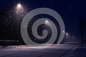 A street lit up by street lamps on the side of the road during a heavy snowfall at night. The street lights illuminate the falling