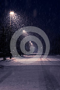 A street lit up by street lamps surrounded by falling snowflakes on the side of the road. The street lights illuminate the heavy