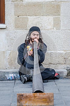 Street musician with long hair and a beard plays long pipe music leaning back against the wall of the house, sitting on the paveme