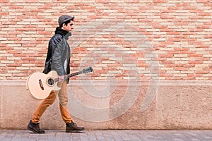 Street musician walking with acoustic guitar. Young handsom man wearing coat and hat outdoors