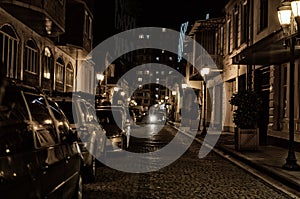 Street of the old night city with paving stone, lit by street lamps with parked cars.