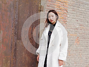 Street photography of a beautiful Chinese woman in white coat with red brick wall and rusty iron door background, looking at you