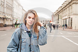 Street portrait of an attractive blonde girl wearing a denim jacket, standing on the street and looking into the camera. Beautiful