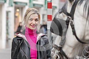 Street portrait of a blonde woman, looking at a horse-drawn carriage in a European city. Concept: tourism and travel,