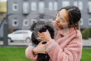 Street portrait of a cute girl with a black little dog in her hands, woman wearing a pink jacket, looking at a pet and smiling.