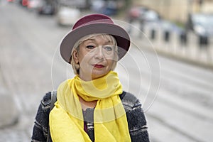 Street portrait of an elderly woman in a hat and yellow scarf against a European urban landscape, selective focus. Concept: excell