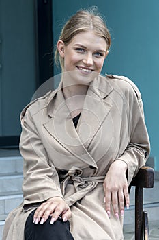 Street portrait of a happy young and beautiful blonde girl 20-25 years old in a light raincoat, sitting on a chair on the street.