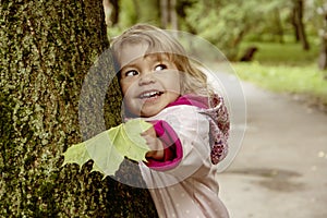 Street portrait of a laughing little girl holding an autumn maple leaf in her hand and hugging a tree. Concept: unity with nature,