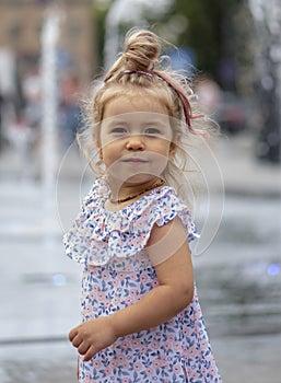 Street portrait of a little blonde girl against the background of city fountains. Her long hair is tied in a ponytail over her hea