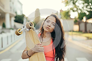 Street portrait of a mulatto girl holding a longboard in her hands, looking in camera, walking with a board in the evening. Girl