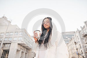 Street portrait of a stylish girl in casual clothing,standing on the street with a cup of coffee in his hand, looks camera.Woman
