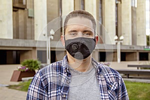 Street portrait of a young man 20-25 years old in a black medical mask on the background of modern city buildings.