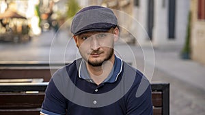 Street portrait of a young man 30 -35 years old in a cap with a beard, sitting on a bench and looking directly into the camera on