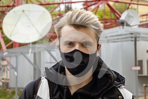 Street portrait of a young man in a black medical mask against the background of telecommunications buildings and satellite dishes