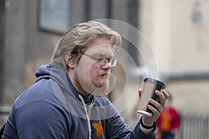 Street portrait of a young man with glasses and a beard 25-30 years old with blond hair drinking coffee from a paper cup against