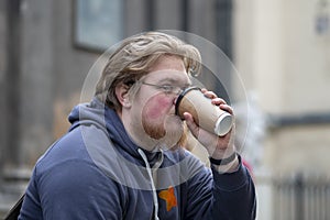 Street portrait of a young man with glasses and a beard 25-30 years old with blond hair drinking coffee from a paper cup against