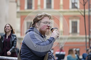 Street portrait of a young man with glasses and a beard 25-30 years old with blond hair drinking coffee from a paper cup against