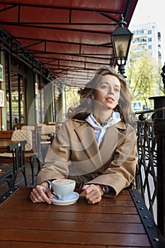 Street portrait of young woman wearing beige coat drinking coffee on cafe veranda with dreamy and thoughtful gaze