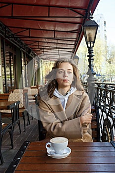 Street portrait of young woman wearing beige coat drinking coffee on cafe veranda with dreamy and thoughtful gaze