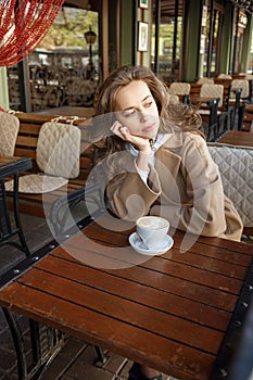 Street portrait of young woman wearing beige coat drinking coffee on cafe veranda with dreamy and thoughtful gaze
