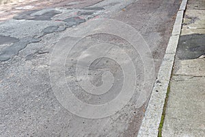 Street and sidewalk in crumbling disrepair, heavily patched asphalt roadway