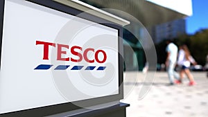 Street signage board with Tesco logo. Blurred office center and walking people background. Editorial 3D rendering