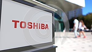 Street signage board with Toshiba Corporation logo. Blurred office center and walking people background. Editorial 3D