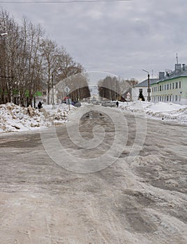A street with a snow-covered road and potholes. Drifts of dirty snow on the roadsides. Houses, road signs, pedestrians.