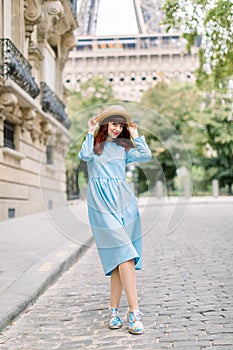 Street view of pretty young red haired lady, wearing elegant blue dress and straw hat, walking during the morning in