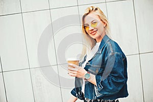 Streetstyle fashion look, pretty cool smiling young woman with coffee cup walking in city, happy female model wearing trendy