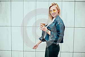 Streetstyle fashion look, pretty cool smiling young woman with coffee cup walking in city, happy female model wearing trendy