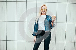 Streetstyle fashion look, pretty cool smiling young woman with coffee cup walking in city, happy female model wearing trendy