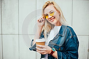 Streetstyle fashion look, pretty cool smiling young woman with coffee cup walking in city, happy female model wearing trendy