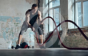 Strength and endurance. Young muscular man doing workout, training with battle ropes indoors. Functional training