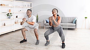 Strength workout concept. Fit black woman and her boyfriend doing squats together at home, panorama