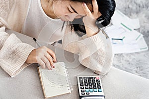 Stress Asian woman calculating her debt having problem with loan looking at list of expenses on notepad with invoices on floor