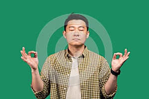 Stress relief concept. Asian man meditating with closed eyes, keeping calm searching for inner balance, green background
