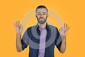 Stress relief concept. Handsome young man meditating with closed eyes, keeping calm on orange studio background