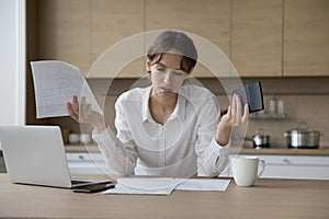 Stressed annoyed young laptop user woman reviewing domestic paper documents