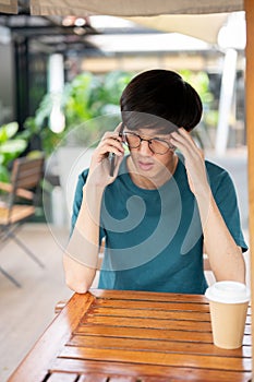 A stressed Asian man talking on the phone while sitting at an outdoor table of a cafe in the city