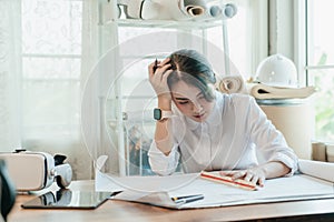 Stressed Asian woman architect having headache while working on blueprint in office
