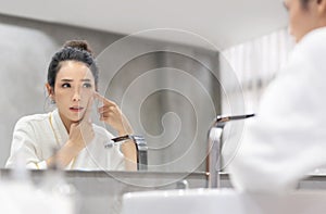 Stressed Asian woman in white Bathrobe looking at pimple on face into the mirror and try to squeeze it