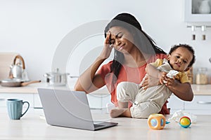 Stressed Black Mother With Baby On Hands Working On Laptop At Home