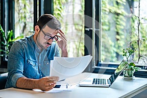 Stressed businessman analyzing documents while working on laptop at home office