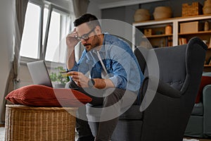 Stressed businessman with credit card paying bills online over laptop while sitting in home office