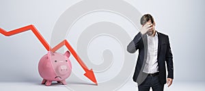 Stressed businessman with falling red chart arrow with piggy bank on white background. Economic recession, losing savings