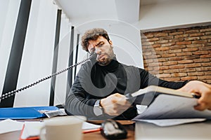 Stressed businessman multitasking with phone and paperwork in a busy office setting, showing frustration and workload