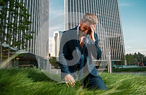 Stressed businessman sitting on grass outside office after project failure. Confused young man frustrated by business