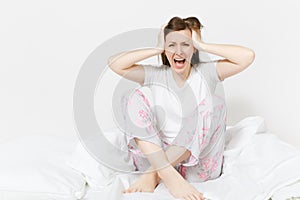 Stressed crazy mad young screaming woman sitting in bed with white sheet, pillow, blanket. Shocked female cover ears