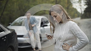 Stressed desperate young woman looking at crashed car with blurred man examining damage at the background. Hopeless